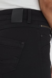 Jeans de mezclilla negra con un parche de cuero con marca, bolsillo con cremallera y remaches de metal. Textura suave y diseño contemporáneo.