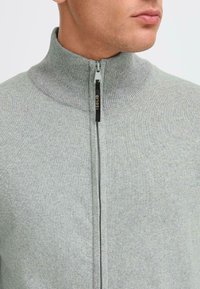 Suéter de cremallera gris claro con un alto cuello, textura acanalada y un tirador negro de la cremallera que lleva la palabra "SOLID."
