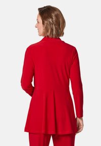 Rood, langemouwen top met een uitlopende zoom en een gladde textuur, met een hoge halslijn en subtiele stikdetails op de achterkant.
