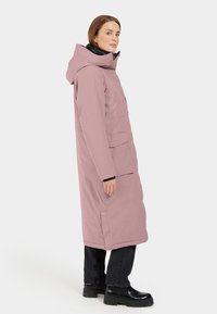 Parka lunga rosa con cappuccio, dotata di tasche laterali doppie e finitura opaca, indossata con pantaloni neri e stivali neri massicci.