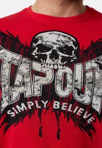 Sarkans kokvilnas T-krekls ar lielu melnu skeletu grafiku un "TAPOUT" tekstu nolietotā baltā fontā, ar traipu detaļām ap to.