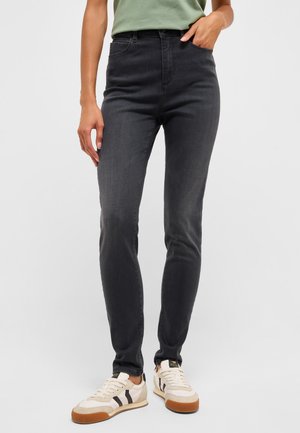 Person wearing fitted dark gray jeans, pale green shirt tucked in, and white sneakers with black stripes and gum soles, standing on white background.