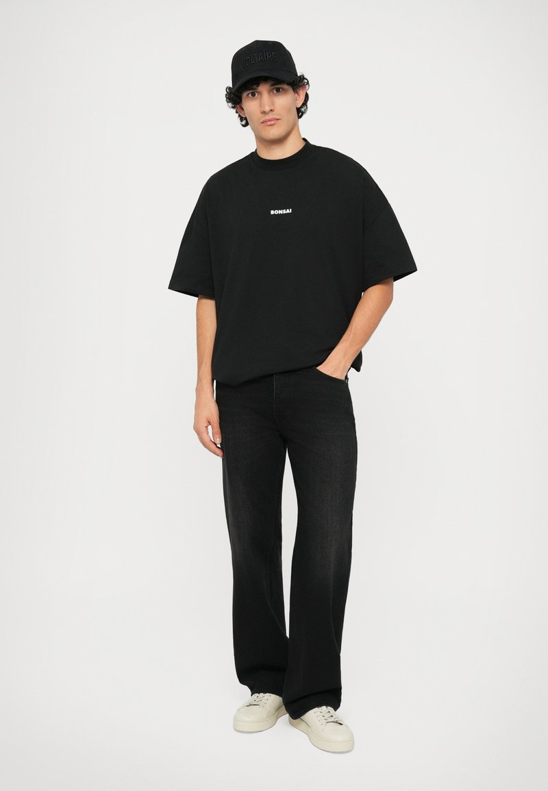 Zwarte katoenen oversized t-shirt met "BONSAI" prints, gecombineerd met zwarte denim broeken en witte sneakers. Inclusief een zwarte pet met "VOLTAIRE" versiering.