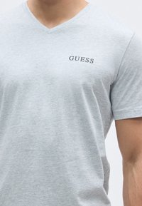 Grå kortærmet V-hals T-shirt lavet af blødt stof, med en lille sort "GUESS"-logo på brystet. Glat tekstur og tætsiddende design.