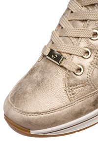 Close-up van een beige sneaker met textielstof met structuur, dikke gevlochten veters, gouden veteroogjes en een metalen veterlabel op een witte zool met een bruine buitenzool.