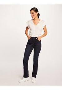 Donkerblauwe hoge taille jeans met witte stiksels en twee voorzakken, gecombineerd met een lichtgrijze V-hals T-shirt en witte sneakers.