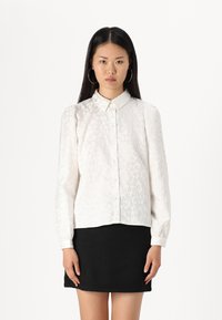 White long-sleeve shirt with collared neckline, textured fabric featuring subtle patterns, button closure, paired with a black textured skirt.