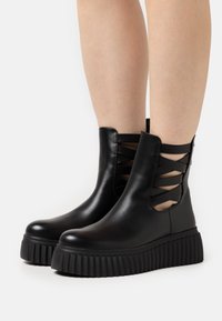 Czarne botki do kostki wykonane z gładkiej skóry, z bocznymi wycięciami i krzyżującymi się paskami. Posiadają grubą, żebrowaną podeszwę для zwiększenia wysokości.