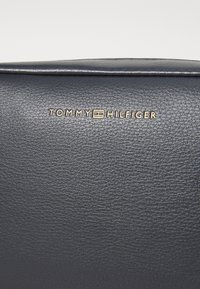 Tamno plava kožna torba s teksturiranom površinom i zlatnim utisnutim logotipom "TOMMY HILFIGER" na prednjoj strani.