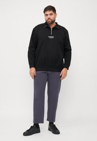 Sudadera negra con cremallera y logo bordado; combinada con pantalones grises y zapatillas negras. El material parece suave y tiene un ajuste relajado.