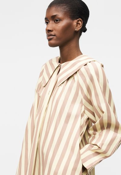 Γυναίκα με σκούρο δέρμα και μαλλιά σε χαμηλό κότσο φοράει μπεζ και λευκό κάθετα ριγέ μπλούζα με φαρδιά γιακά, κοιτάζοντας πλάγια σε μονόχρωμο φόντο.