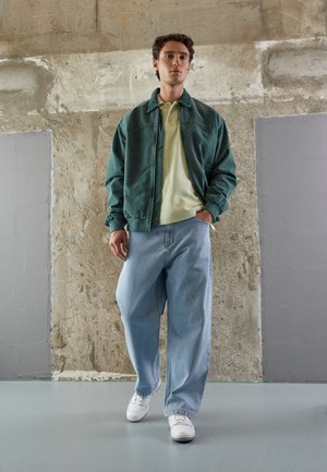 Green bomber jacket over a light yellow polo, paired with loose blue jeans and white sneakers. Smooth fabrics, relaxed fit, minimal hardware.