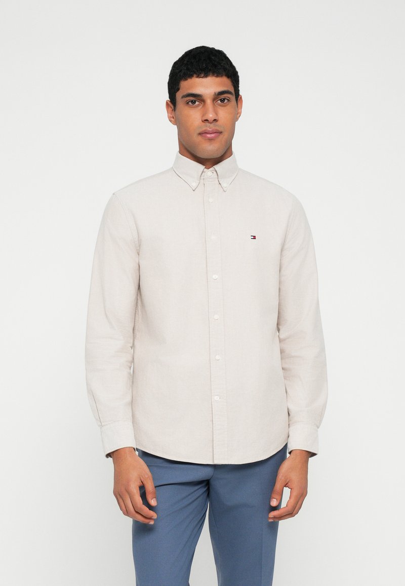 Svetlo béžová košeľa na gombíky s golierom, dlhými rukávmi a jemnou textúrou. Na ľavej strane na hrudi je malá logo.