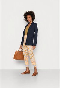 Mulher com cabelo encaracolado, vestindo um blazer azul-marinho, uma blusa mostarda, calças floridas e mocassins castanhos, segurando uma grande bolsa castanha, de pé contra um fundo branco.