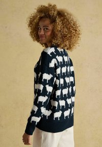 Frau mit lockigem Haar in einem schwarzen Pullover mit weißen Schafen, die seitlich vor einem schlichten beigen Hintergrund steht.