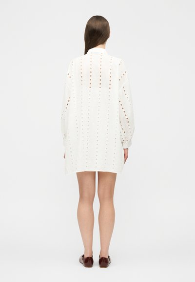 Kvinde med langt brunt hår iført en hvid, langærmet minikjole med hulmønster og brune flade sko, stående med ryggen til mod en hvid baggrund.