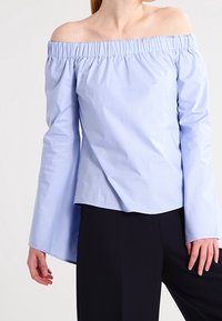 Jasnoniebieska bluzka typu off-the-shoulder z marszczonym dekoltem, długimi rękawami i luźnym krojem; wykonana z gładkiego materiału o lekkim połysku.