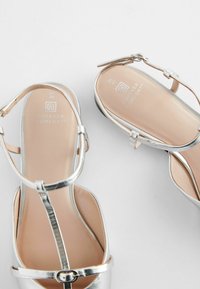 Silverfärgade remsandaler med metallisk finish, öppen tå-design och ankelremmar. Mjuka tan-färgade innersulor med "Forever Comfort"-logotyp.