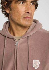 Sweat à capuche en polaire de couleur mauve doux avec une fermeture éclair à l'avant, des cordons de serrage et un logo brodé blanc sur la poitrine gauche. Texture lisse.