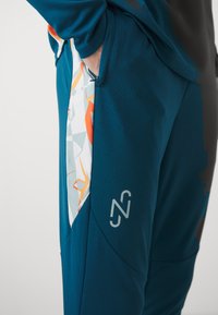 Teal sportieve broek met een gestructureerd oppervlak, voorzien van een kleurrijk geometrisch patroon aan de zijkant, een subtiele ritszak en logo-detail.