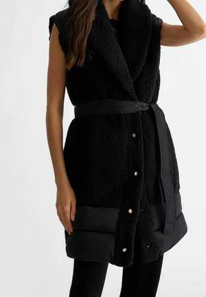 Bezrekawnik w czarnym kolorze z fakturą, z guzikami i paskiem, noszony na czarnych legginsach na prostym białym tle.