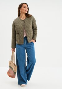 Gepolsterte olivgrüne Jacke mit Rundhalsausschnitt und Druckknöpfen, kombiniert mit hellblauen weit geschnittenen Jeans. Das Model hält eine mehrfarbige gewebte Tragetasche.