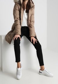 Béžová péřová bunda s kapucí podšitou kožešinou, kombinovaná s černými úzkými kalhotami a bílými teniskami s kovovými očky a červenými akcenty.