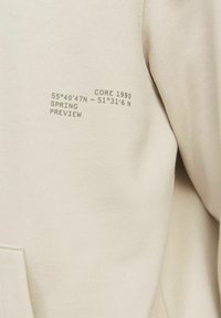 Light beige sweatshirt with small dark text on chest reading "CORE 1990 55°40'47N - 51°31'6N SPRING PREVIEW".