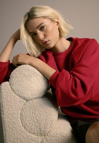 Punainen collegepaita, jossa on pyöreä kaulus ja löysät hihat, yhdistettynä teksturoituun beigeen tuoliin, jossa on pyöreitä, pehmeitä yksityiskohtia.