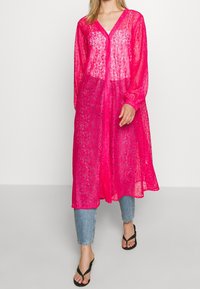 Abito lungo rosa brillante e trasparente, con maniche lunghe e scollatura a V, caratterizzato da un'apertura con bottoni sul davanti e una silhouette fluida, indossato sopra dei jeans azzurri chiari.