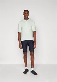 Svetlomätový krátky rukáv s polovičným zipsom na krku v kombinácii s tmavomodrými cyklistickými šortkami. Modelka má na sebe čierne športové topánky a biele ponožky.