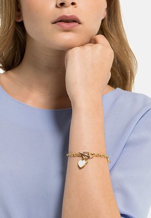 Bracelet doré avec un design de chaîne, présentant un charm en forme de cœur blanc et un fermoir à bascule, porté sur un poignet contre un tissu bleu clair.