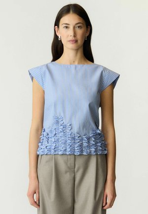 Femme portant un haut sans manches rayé bleu et blanc avec des détails de tissu texturé près de l'ourlet et un pantalon beige, debout devant un fond uni.