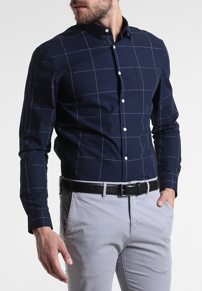Cămașă cu mânecă lungă de culoare albastră închis, cu un model de grilaj, având linii albe, cu închidere prin butoni și guler clasic. Asortată cu pantaloni gri deschis.