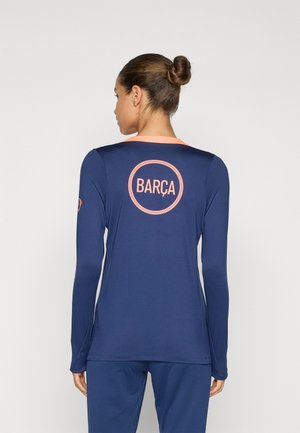 Námořnická modrá košile s dlouhým rukávem a růžovými akcenty, se zaobleným logem "BARÇA" na zádech. Hladký materiál a sportovní střih.