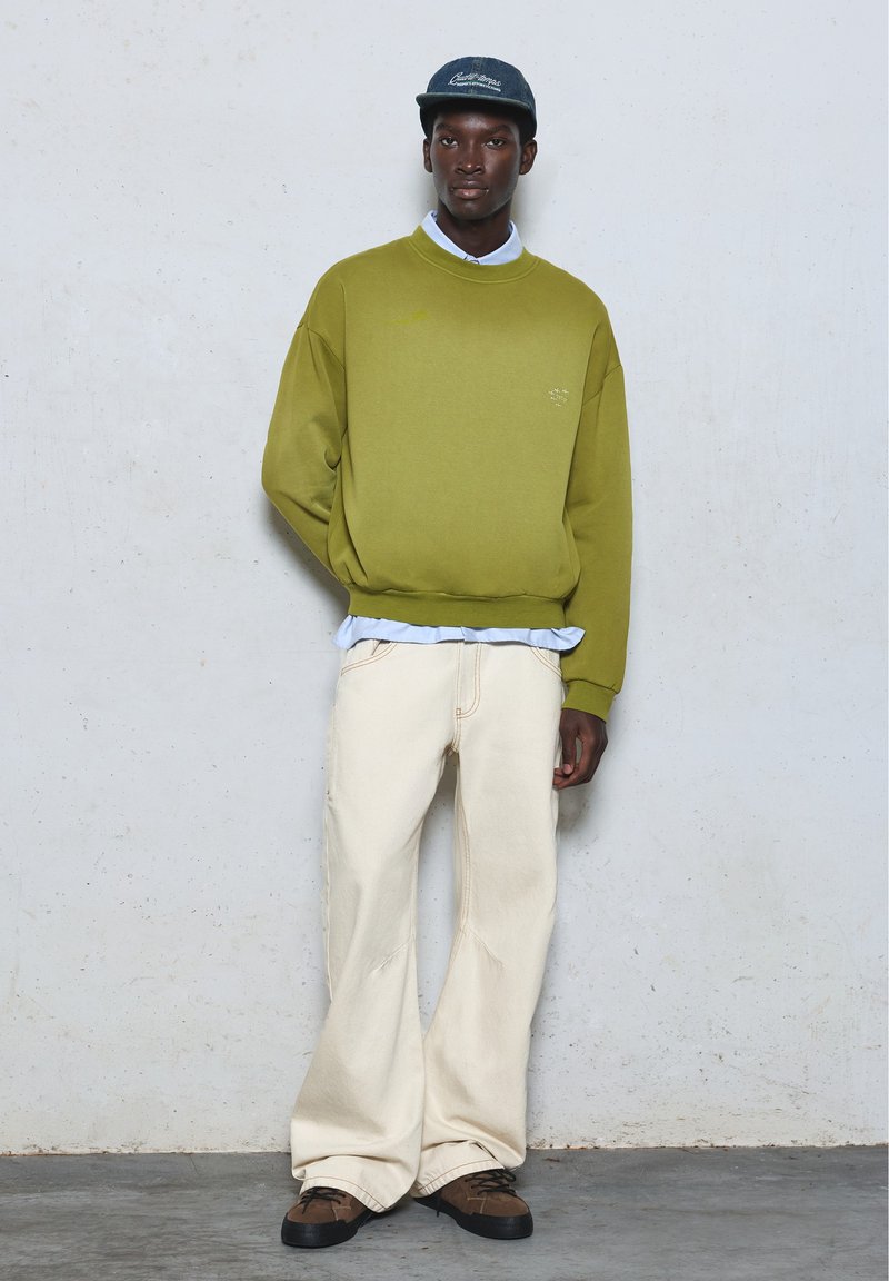 Un jeune homme se tient contre un mur blanc, portant un sweat-shirt vert olive, un pantalon blanc, des chaussures marron et une casquette sombre.