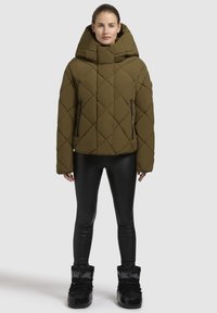 Olijfgroene gewatteerde pufferjack met een hoge kraag en grote capuchon. Bevat ritszakken aan de zijkanten en heeft een boxy silhouet. Gecombineerd met zwarte leggings en laarzen.