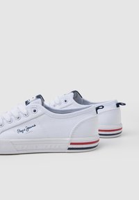 Sneakers bianchi in tela con design a taglio basso, dotati di un logo blu, strisce rosse e blu navy sulla suola e chiusura con lacci.