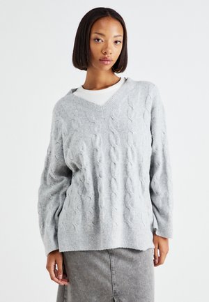 Suéter gris de punto trenzado con escote en V y mangas largas, con patrones texturizados y un dobladillo acanalado, llevado sobre una blusa blanca.