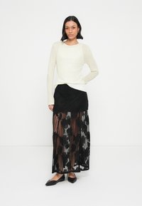 Γυναίκα που στέκεται φορώντας μια κρεμ μπλούζα με μακριά μανίκια, μαύρη μάξι φούστα από δαντέλα με floral μοτίβα και μαύρα μυτερά μπαλαρίνια, μπροστά από λευκό φόντο.