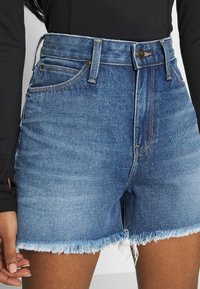 Pantalones cortos de mezclilla en azul claro con dobladillo desflecado, diseño de cintura alta, cinco bolsillos y cierre frontal con botón. Textura deslavada con costuras en contraste.