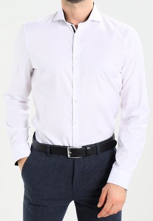 Man wearing a white long-sleeve dress shirt, dark belt, and dark gray trousers, with one hand in pocket against plain background.