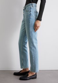 Jeans de tiro alto en azul claro con corte recto, que presentan una textura deslavada, herrajes metálicos dorados y bolsillos laterales. Zapatos negros tipo mocasín.