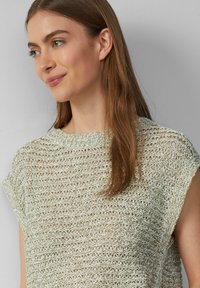 Jasnozielony sweter z dzianiny o luźnym, otwartym splocie, okrągłym dekolcie i krótkich rękawach. Tekstura wydaje się przewiewna z subtelnym połyskiem.
