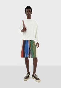 Langärmliges weißes Hemd, kombiniert mit mehrfarbigen gestreiften Shorts in Blau, Grün, Rot und Braun, ergänzt durch braune Sneaker und einen Schultergurt.