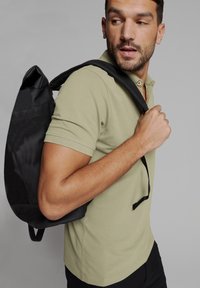 Zwarte rugzak met een slank, gestructureerd ontwerp, gedragen op één schouder over een licht olijfgroene polo met een klassieke kraag.