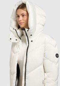 Biała kurtka puchowa z dużym kapturem, pikowaną teksturą i czarnym zamkiem błyskawicznym. Posiada zatrzaski i naszywkę z logo na rękawie.
