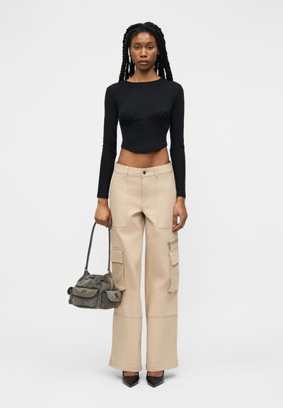 Γυναίκα με μακριές κοτσίδες φοράει μαύρο crop top με μακριά μανίκια, μπεζ παντελόνι cargo με φαρδιά μπατζάκια, μαύρες μυτερές γόβες, κρατώντας γκρι τσάντα στο δεξί χέρι.