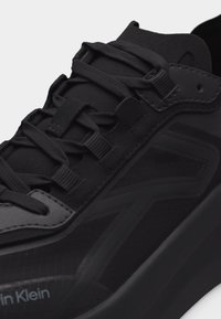 Gros plan sur une basket noire Calvin Klein montrant des panneaux latéraux texturés, des lacets en tissu et des détails de languette rembourrée.