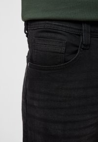 Schwarze Denim-Jeans mit klarem Design, sichtbarer Seitentasche und dezenter Textur. Der Stoff wirkt glatt mit minimalen Details.